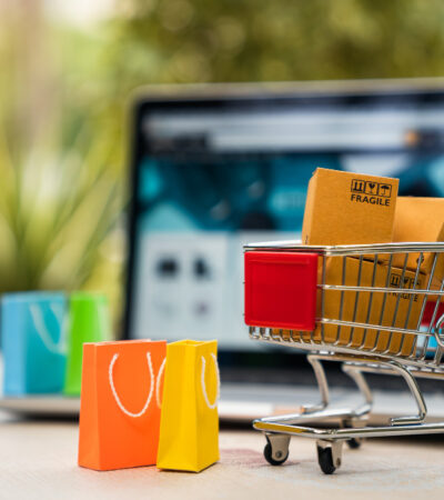 Ilustracja przedstawiająca model wózka sklepowego pełnego zakupów, w tle laptop